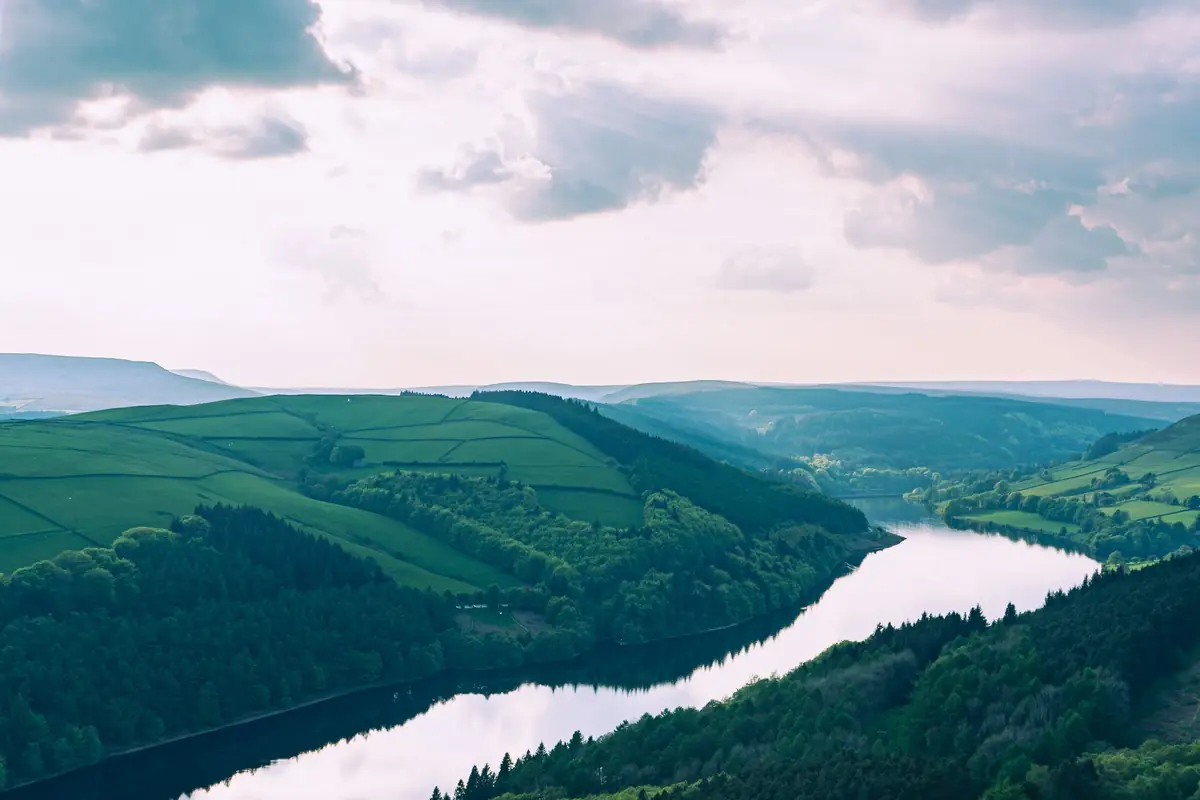A reservoir winding between wooded hills and farmland under a cloudy sky.
